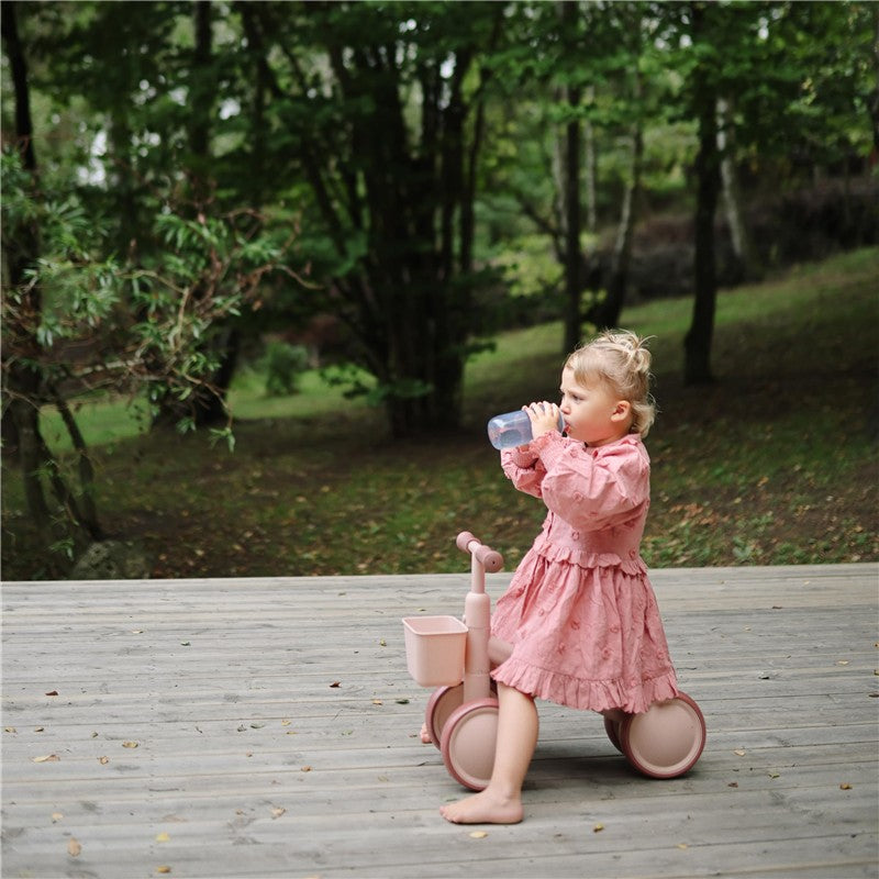 A toddler in a pink dress sits on a small pink bike, drinking from a blue Mushie soft spout sippy cup in a park.