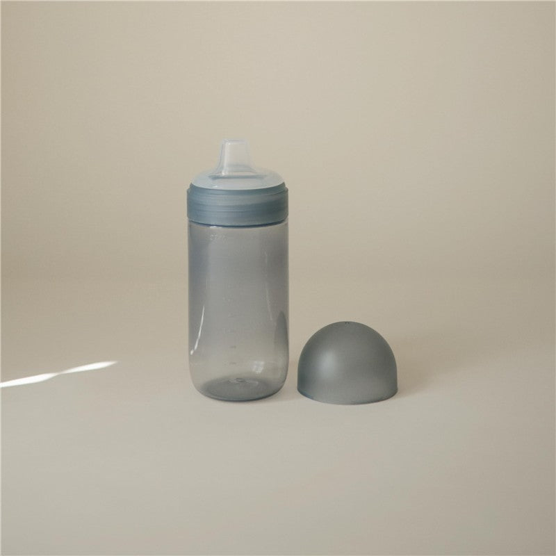 Mushie Soft Spout Sippy Cup in Tradewinds color, featuring a transparent body and a soft spout lid, with a dome cap nearby.
