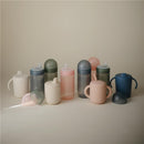 Mushie Soft Spout Sippy Cups in various pastel colors, featuring soft spouts and ergonomic handles for easy grip.