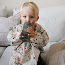 A toddler in a colorful, patterned bib holds a Mushie Soft Spout Sippy Cup in Tradewinds while sitting on a couch.