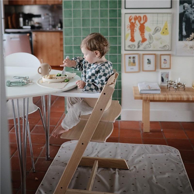 Toddler in a high chair, enjoying a meal on a patterned Mushie Splat Mat in blush color, with a green tiled background.
