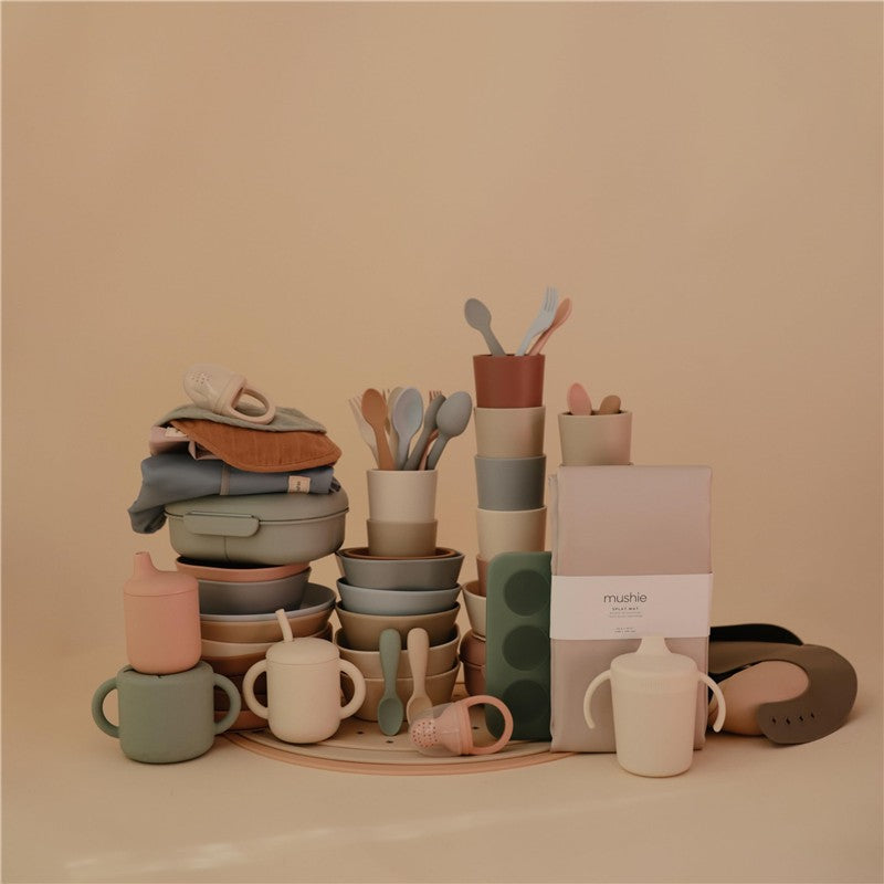 Mushie Splat Mat in Blush surrounded by various coordinated stacking bowls, cups, utensils, and soft goods.