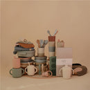 Mushie Splat Mat in Blush surrounded by various coordinated stacking bowls, cups, utensils, and soft goods.