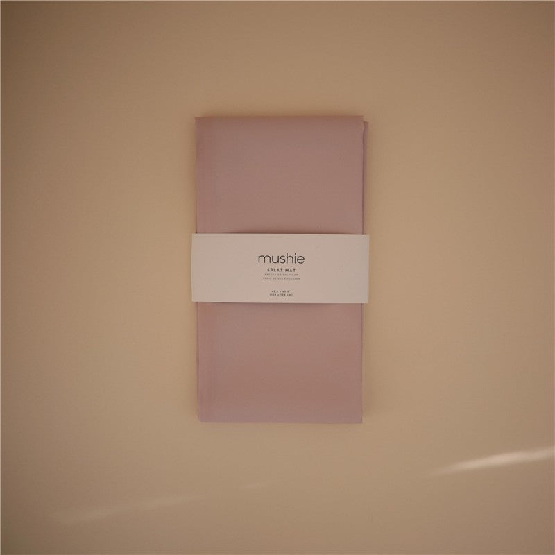 Mushie Splat Mat in blush color, featuring a white band with brand name and product details, laid flat on a neutral background.