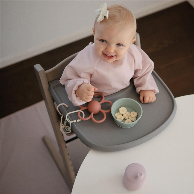 A happy baby in a pink smock sits in a wooden high chair, playing with a silicone toy and eating banana slices from a green bowl.