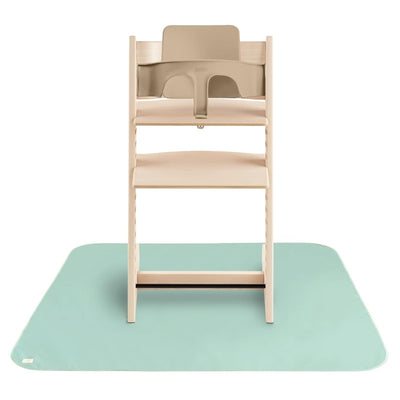 Mushie Splat Mat in Roman Green under a wooden high chair, designed to protect floors during mealtime.