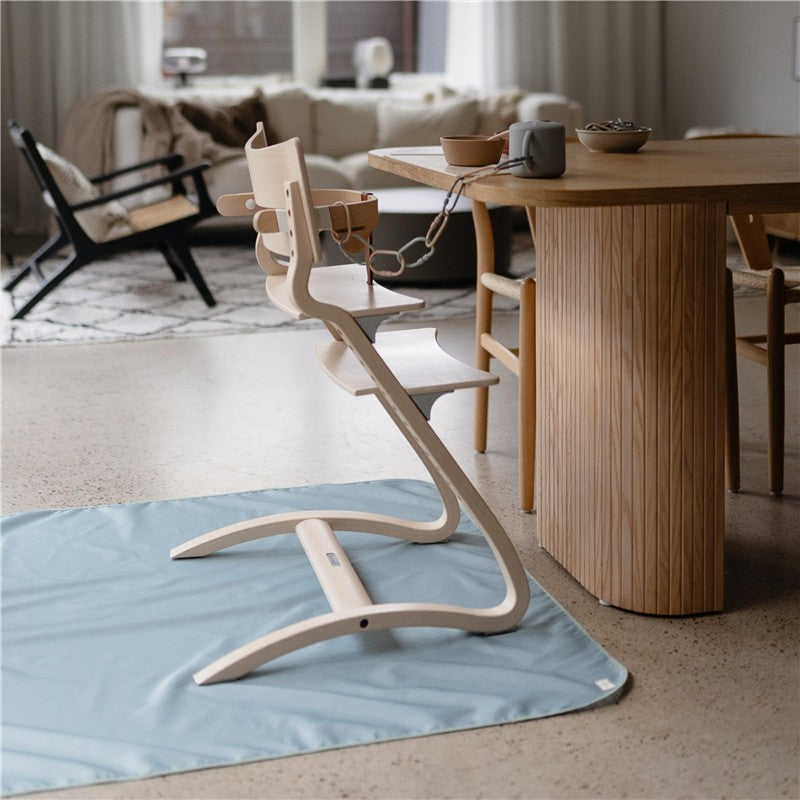 Mushie Splat Mat in Roman Green positioned under a wooden high chair in a modern dining area.