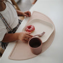 Mushie ivory silicone plate on a pink mat, featuring a donut and a cup, held by a child with a fork.