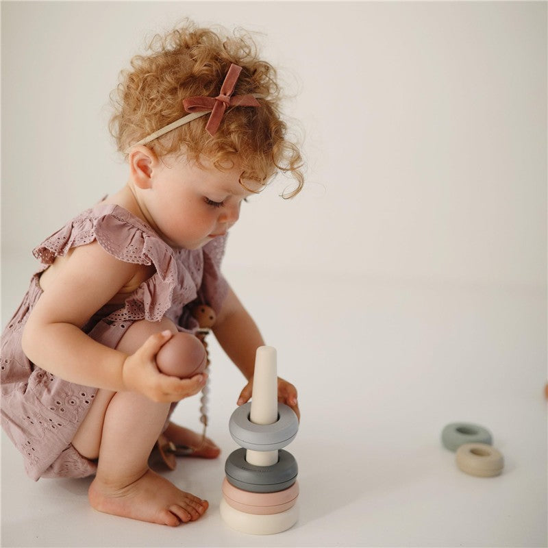 A toddler in a dusty pink outfit plays with Mushie Stacking Rings, a colorful toy with stacked rings.