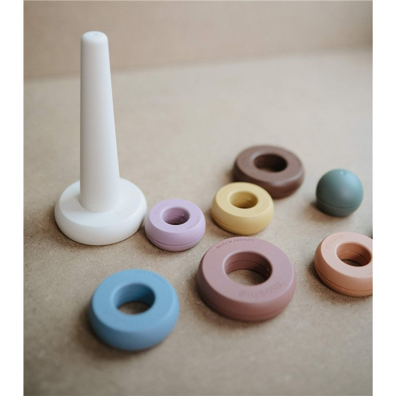 Mushie Stacking Rings set features a white center post and colorful silicone rings in shades of blue, pink, brown, and yellow.