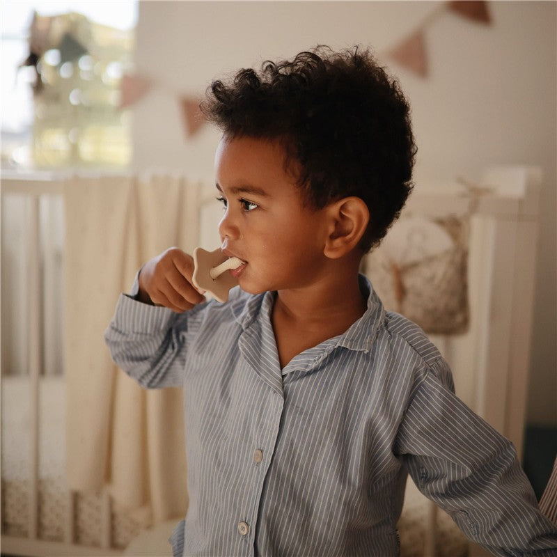 A young child in a striped blue shirt uses a Mushie Silicone Star Training Toothbrush, holding it in their mouth.