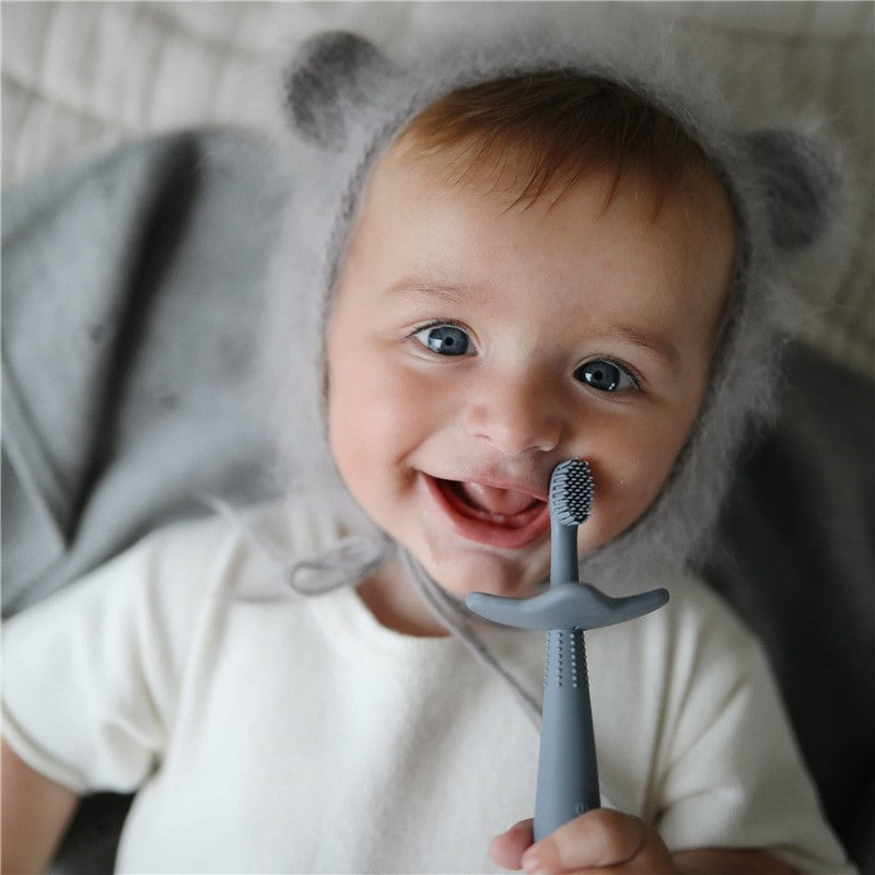 Smiling baby in a gray bear hat holds a Mushie silicone star training toothbrush in Cambridge Blue.