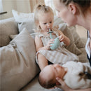 Child using a Cambridge Blue Mushie Silicone Star Training Toothbrush while seated on a couch beside a caregiver.
