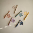 Mushie silicone training toothbrushes in pastel colors surrounded by flower-shaped bases on a neutral background.