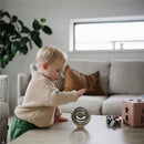 A toddler in a beige sweater plays with a Mushie Suction Spinner Ball on a wooden table, surrounded by colorful toys.