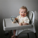 Smiling baby in a high chair holding a Mushie Suction Spinner Ball toy, wearing a cream-colored shirt with heart patterns.