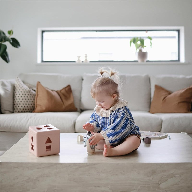 Toddler playing with the Mushie Suction Spinner Ball on a coffee table, surrounded by soft toys and a colorful block.