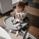 A toddler in a high chair plays with a Mushie Sunshine Suction Spinner Toy and enjoys a bowl of cereal.