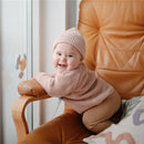 A smiling baby wearing a pink sweater and hat sits on a brown chair, reaching towards a colorful suction spinner toy on the window.