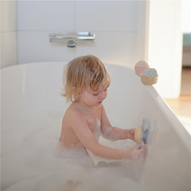 A toddler bathes in a bubble-filled tub, playing with the Mushie Sunshine Suction Spinner Toy attached to the side.