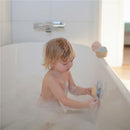 A toddler bathes in a bubble-filled tub, playing with the Mushie Sunshine Suction Spinner Toy attached to the side.