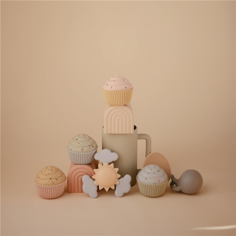 Colorful silicone toys including a sun, clouds, and cupcakes arranged around a beige cup, designed for sensory play.