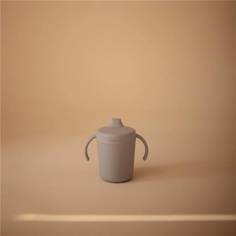 Gray Mushie Trainer Sippy Cup with two handles and a spill-proof top, set against a soft beige background.