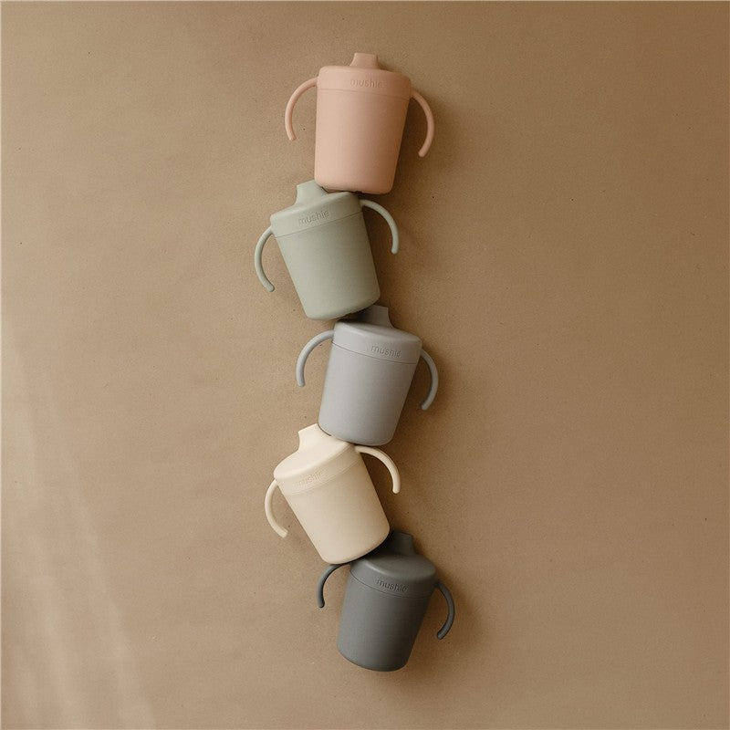 Mushie Trainer Sippy Cups in pastel colors stacked vertically on a neutral background, featuring easy-to-hold handles.