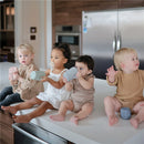 Four toddlers sit on a kitchen countertop holding Mushie Trainer Sippy Cups in pastel colors, showcasing their playful moments together.