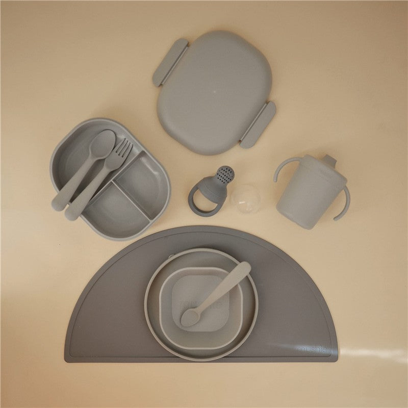 Mushie Trainer Sippy Cup in Cloud color alongside a gray plate, utensils, and a spill-proof lid on a light surface.