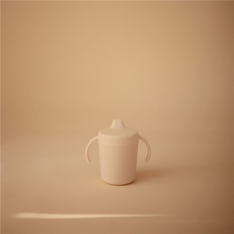 Ivory Mushie Trainer Sippy Cup with ergonomic handles and a lid, designed for easy drinking for toddlers.