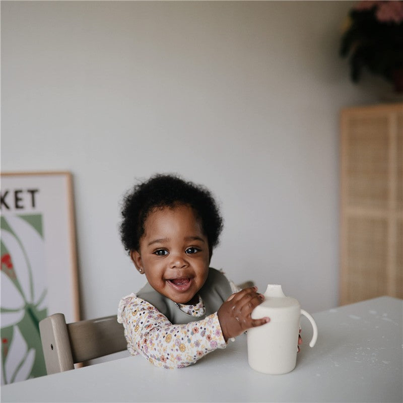 Smiling baby in a high chair holding a Mushie Trainer Sippy Cup in ivory, wearing a floral long-sleeve top.