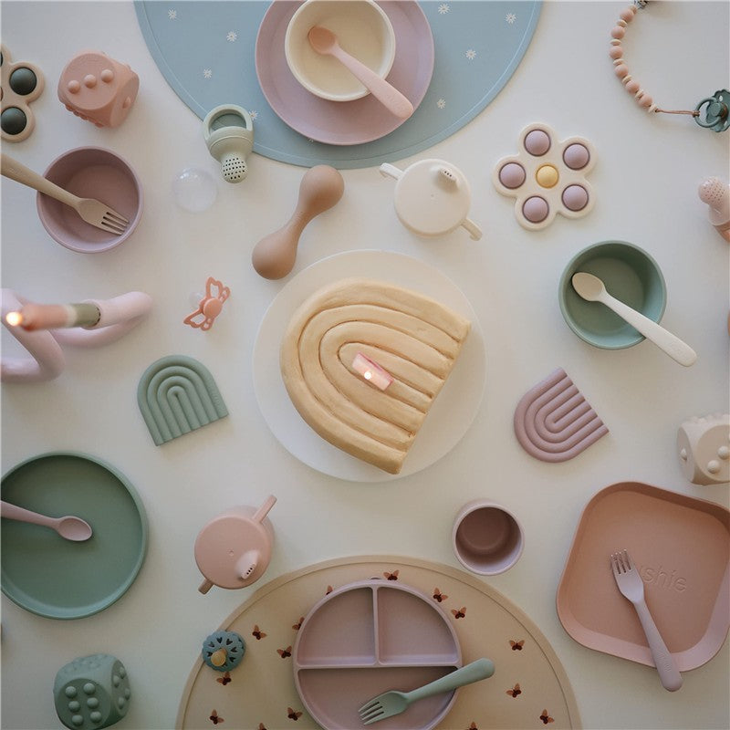 Trainer sippy cup in ivory surrounded by plates, utensils, and a toy cake with a candle on a light surface.