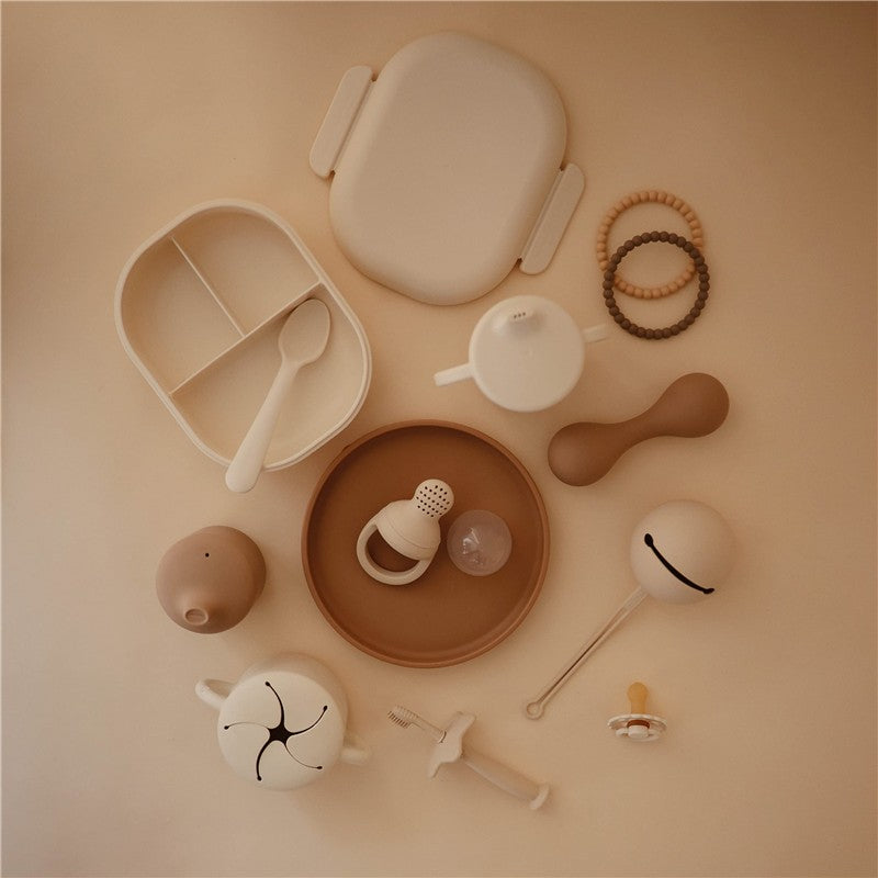 Mushie Trainer Sippy Cup in ivory surrounded by various soft feeding and teething accessories on a neutral background.