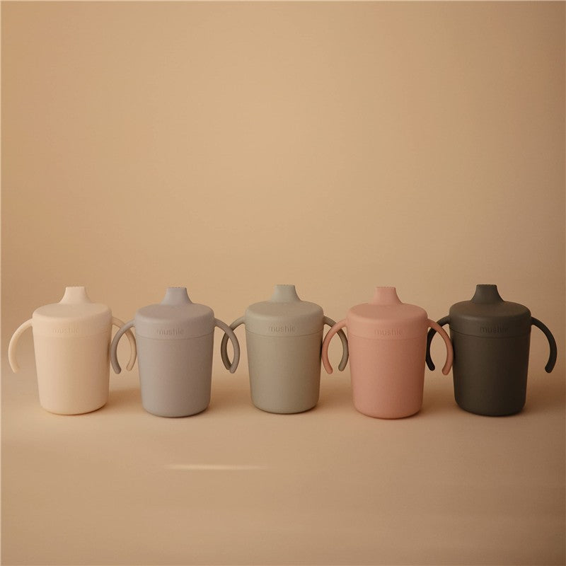 Trainer sippy cups in ivory, grey, green, pink, and dark grey arranged in a row, featuring two easy-to-grip handles.