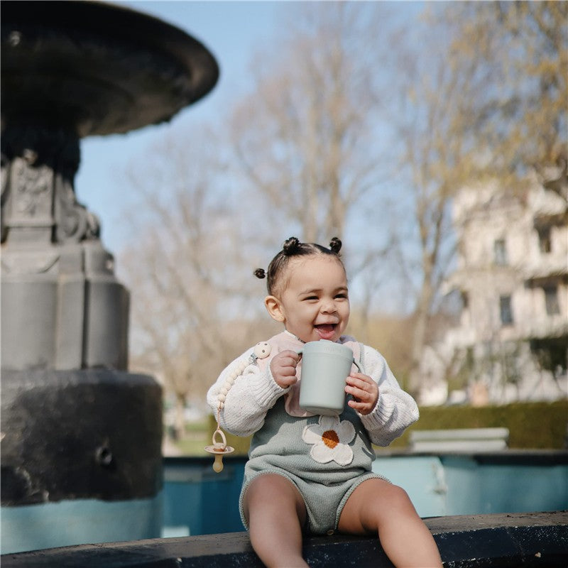 Smiling toddler in a sage green outfit holding a Mushie Trainer Sippy Cup, sitting by a fountain outdoors.