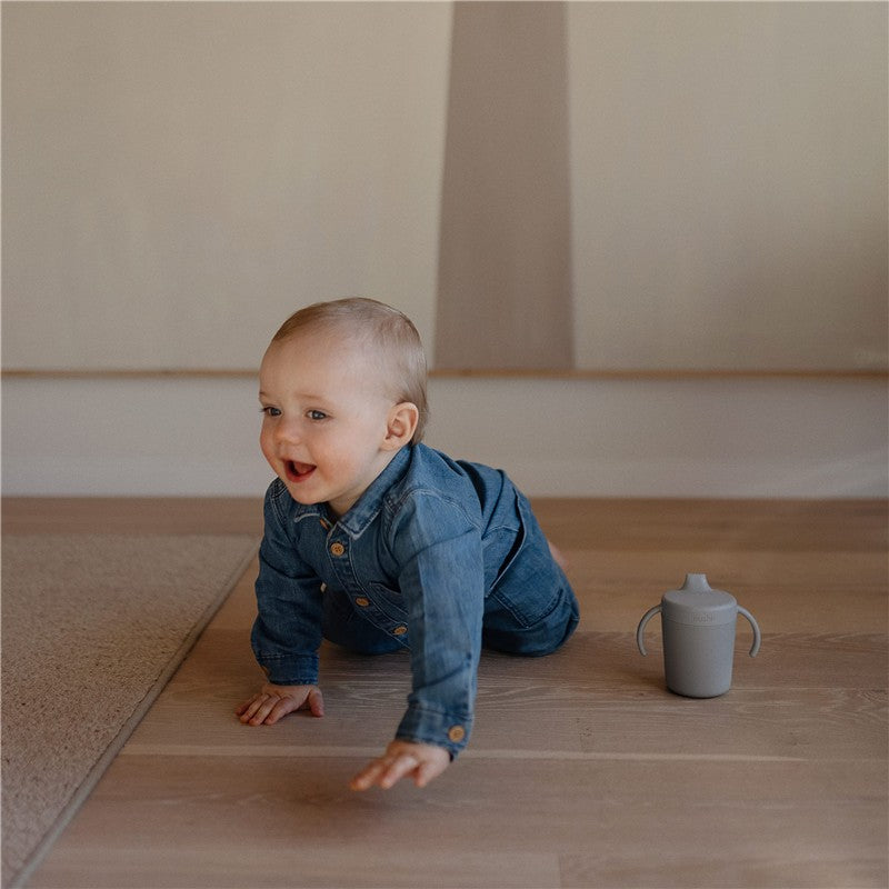 Baby crawling on the floor, wearing a denim outfit, next to a gray Mushie Trainer Sippy Cup.