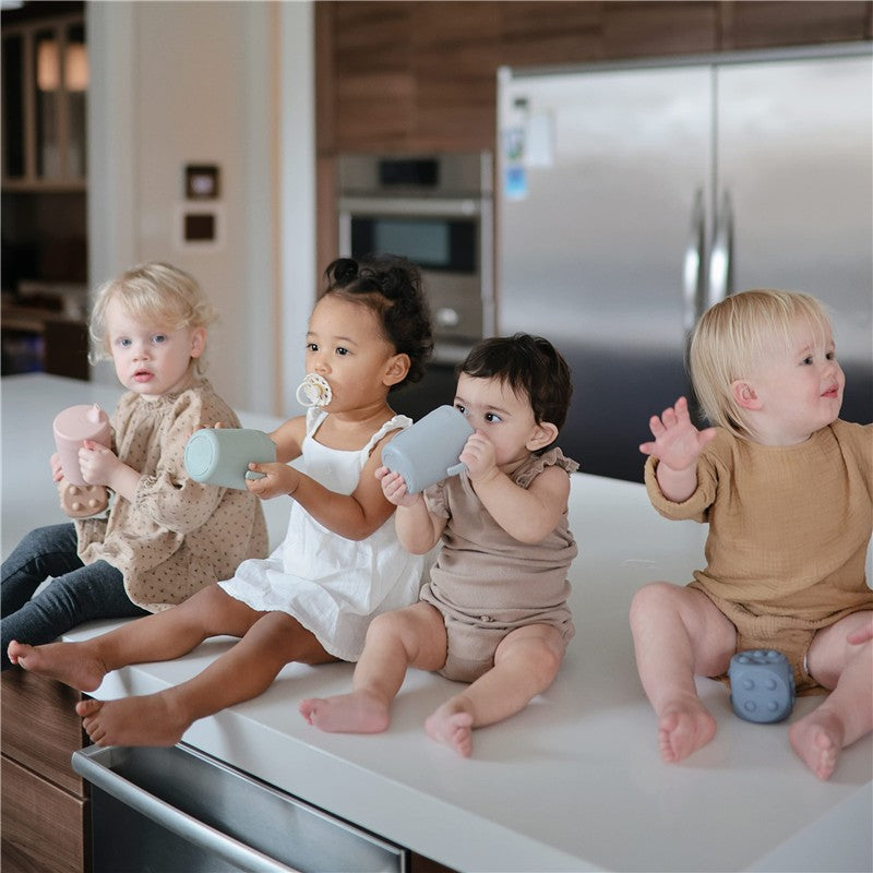 Four toddlers sit on a kitchen counter with sippy cups. One holds a pink cup, two have blue and green cups, and one has a pacifier.