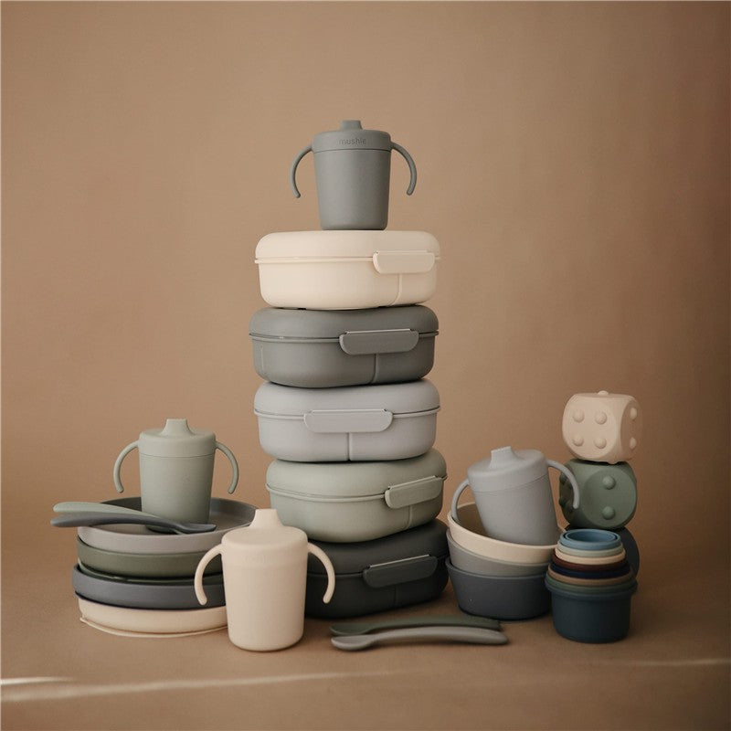 Mushie Trainer Sippy Cup in smoke color, surrounded by various stackable silicone containers and utensils in muted tones.