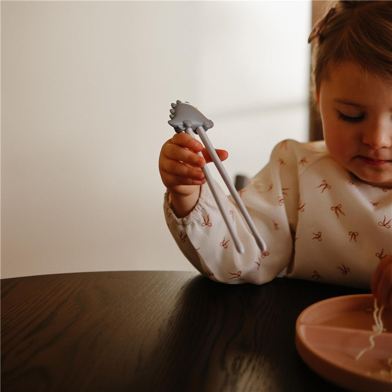 Child holding gray dinosaur-shaped training chopsticks while practicing picking up food from a pink plate.