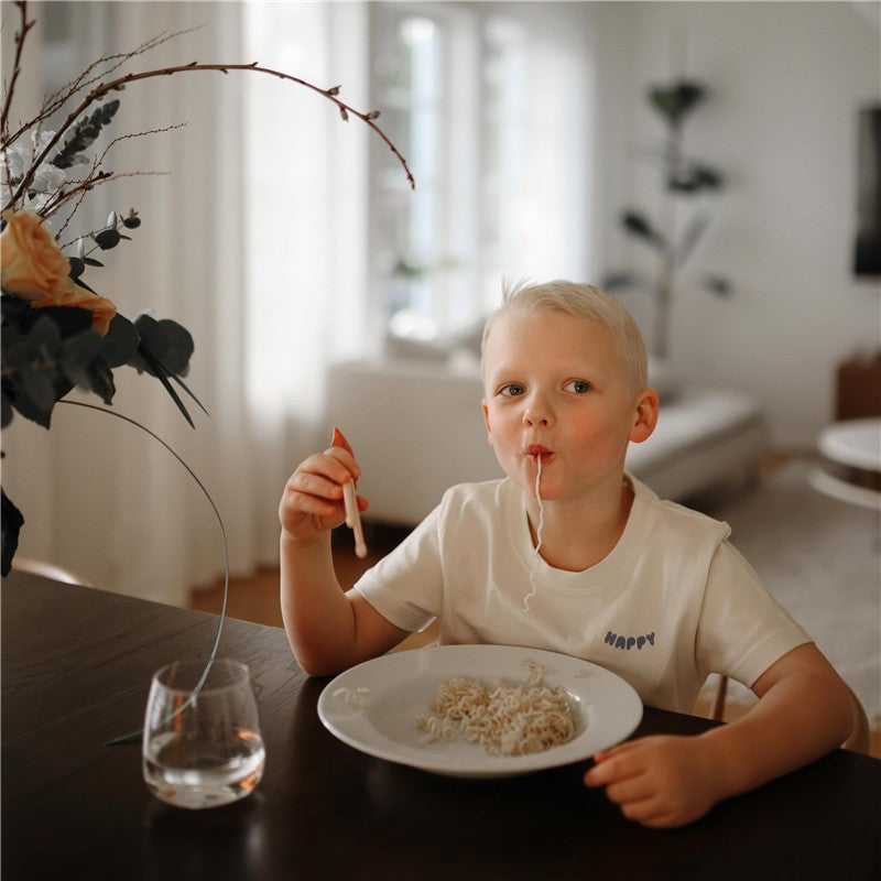 Child using pink training chopsticks to eat noodles from a white plate, smiling at the camera in a cozy indoor setting.