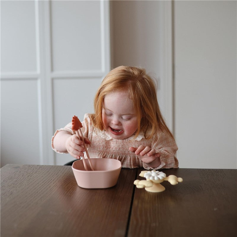 A child uses Mushie Training Chopsticks in a pink bowl, focused and enjoying the activity at a wooden table.