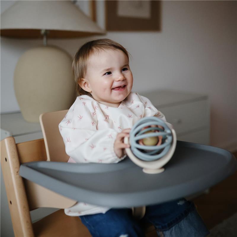 Baby smiling while seated in a high chair, wearing a Mushie water-resistant long sleeve bib with bows, holding a toy.