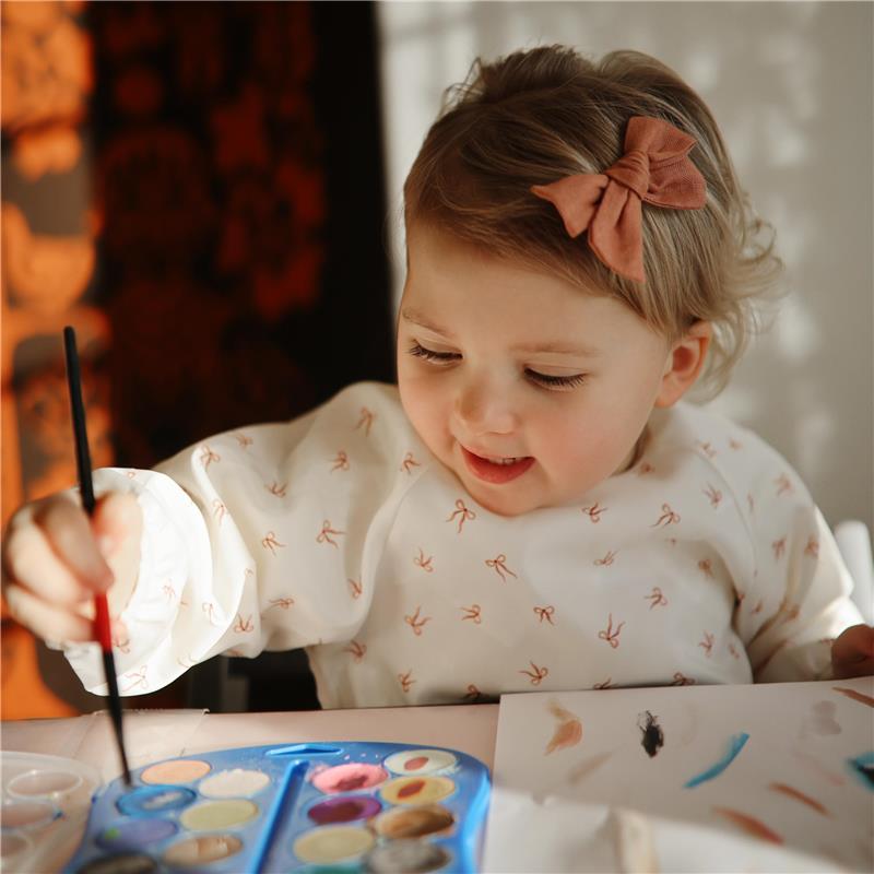Toddler wearing a water-resistant bib with bows, happily painting on paper with a brush and watercolor palette.