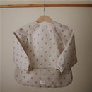 Water-resistant long sleeve baby bib with cherries pattern, featuring adjustable fit and elastic cuffs, by Mushie.