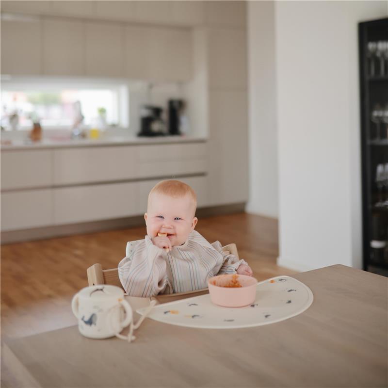 Baby seated in a high chair wearing a striped long sleeve bib, smiling and eating from a pink bowl, with a dining area in the background.