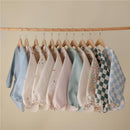 A variety of long sleeve baby bibs in different colors and patterns are hung on wooden hangers.