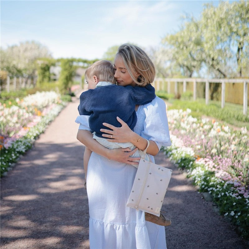 Mother holding a young child in a flower garden, with a Mushie Water Resistant Wet Bag featuring a bee pattern visible.
