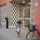 Child in a denim dress holding a Mushie water-resistant wet bag outside a building with a bicycle nearby.