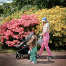 A woman in pink overalls pushes a stroller with a child while holding a bag, accompanied by another child in green overalls, surrounded by colorful flowers.